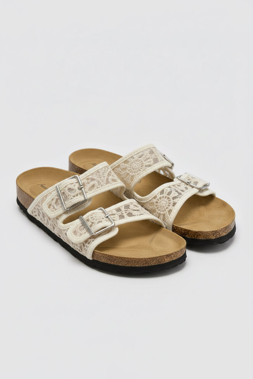 A pair of women's slide sandals with beige lace straps, silver buckles, and a brown cork-look midsole. 