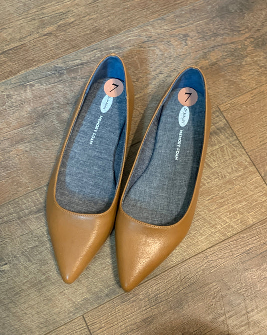 A pair of tan pointed-toe flats with grayish blue fabric footbeds, shown on a wood floor. The insoles show the brand name "Dr. Scholls and Memory Foam'.